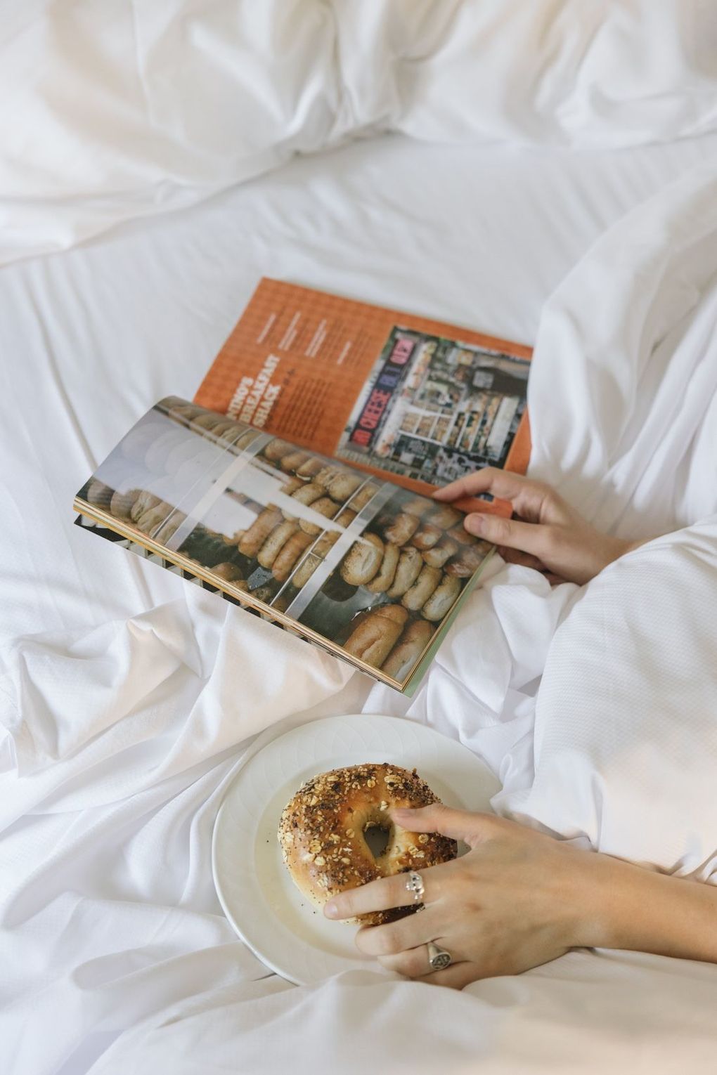 Person in bed, reading a magazine and holding a bagel on a plate.