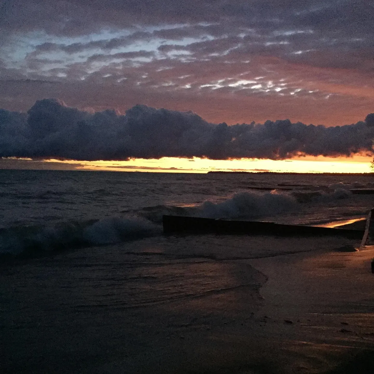 Dark ocean waves at sunset under a cloudy purple sky, with a glowing orange horizon.