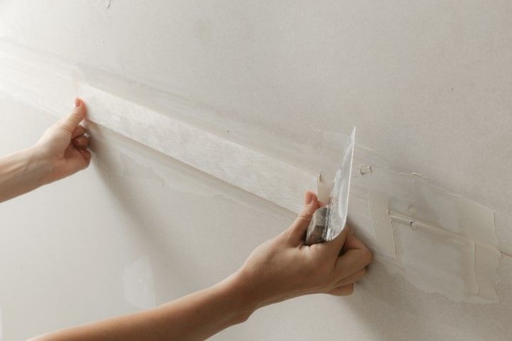 Hands applying painter’s tape along a white wall seam with a putty knife.