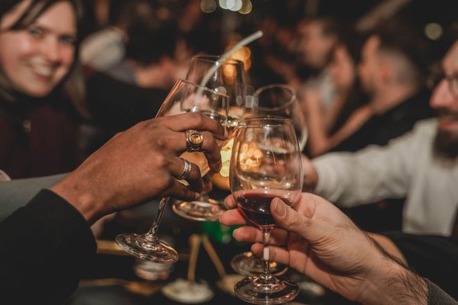 Des personnes trinquent dans un bar ; mains levées, verres qui tintent. Visages flous en arrière-plan.