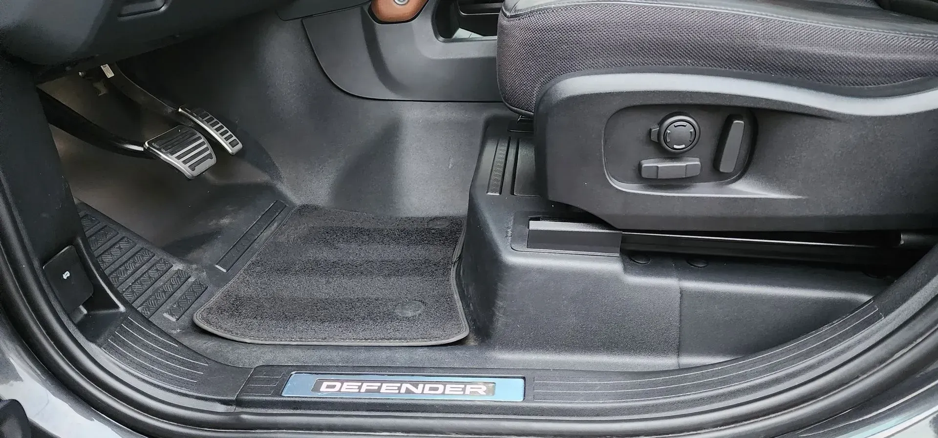 Car door sill and footwell with black floor mat and seat adjustment lever.