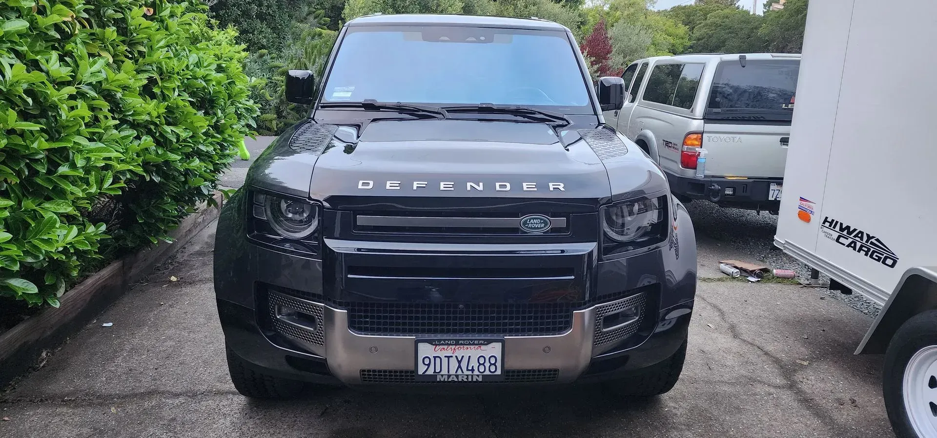 Black Porsche SUV parked in a narrow driveway beside a white van and hedge rows