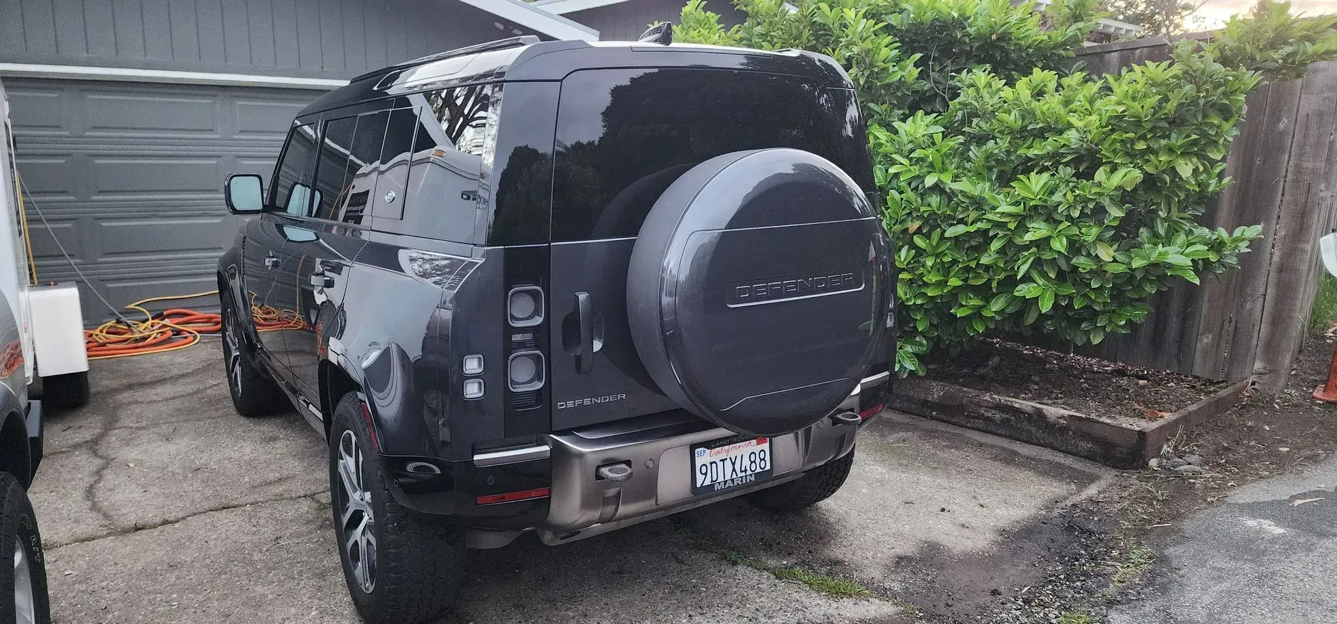 Black SUV parked in a driveway beside a garage, rear view with spare tire cover and wet pavement