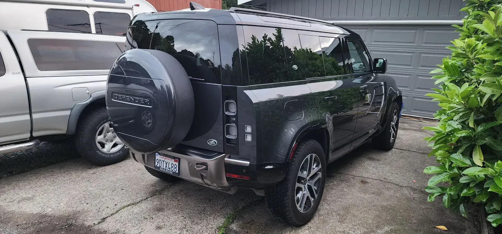 Black SUV parked in a driveway beside a silver truck, rear view with spare tire cover and wet pavement