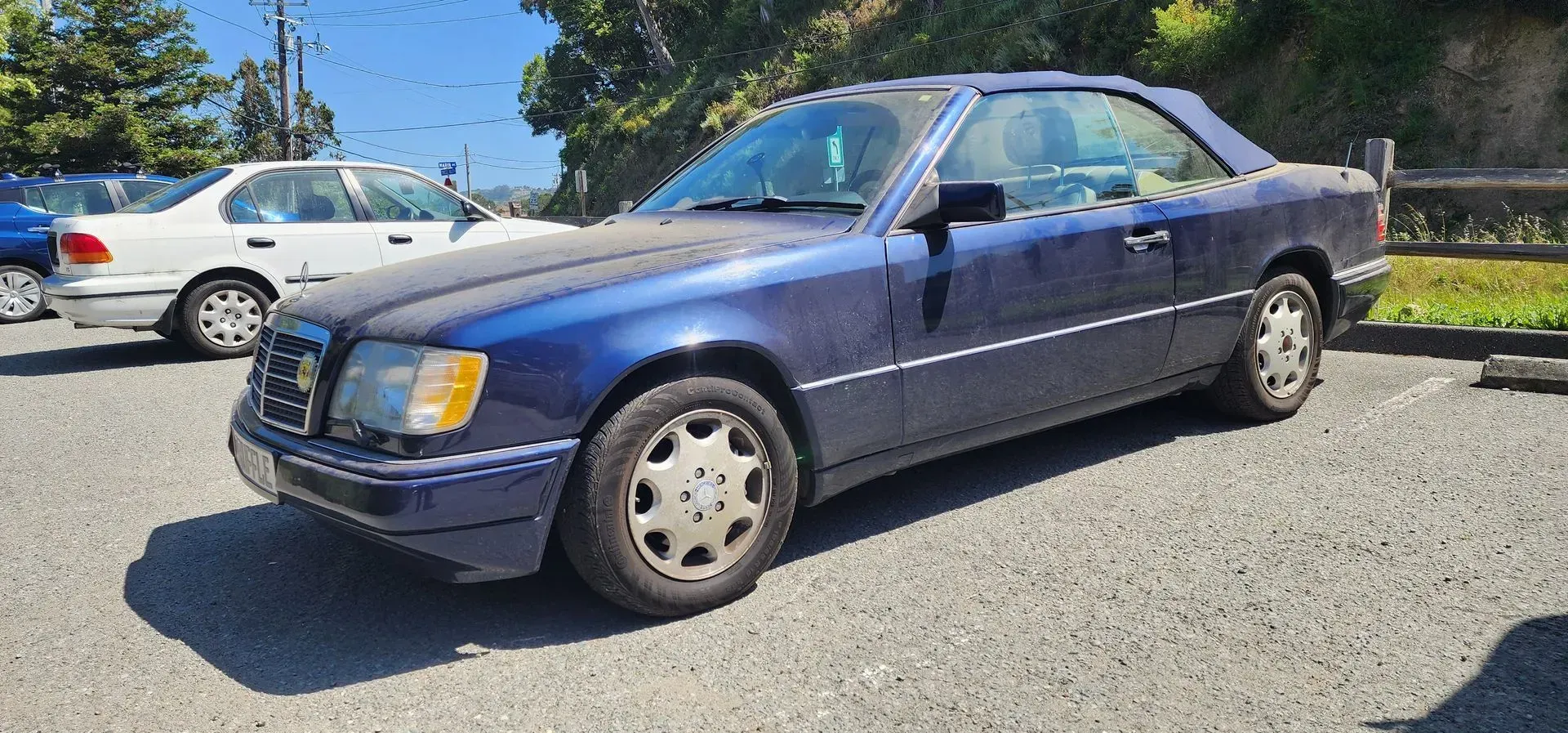 Blue sedan parked on a sunny gravel lot, with other cars and trees in the background