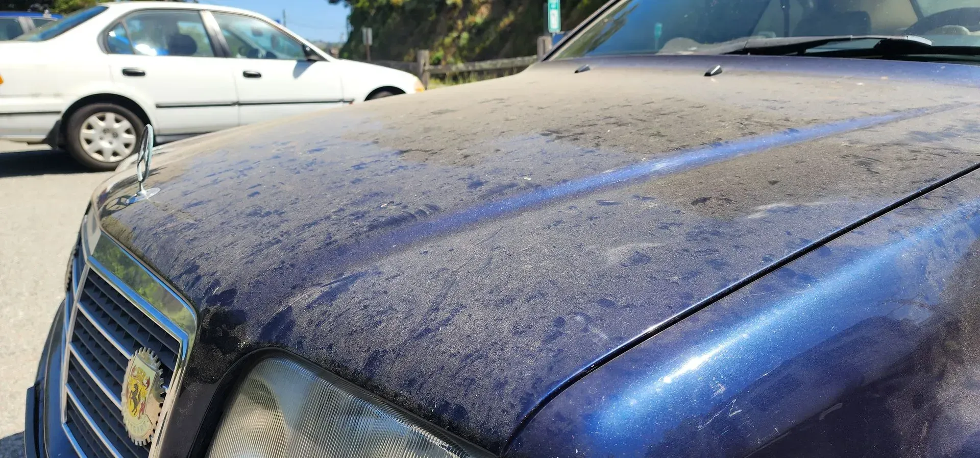Close-up of a dusty blue car hood and windshield with a white car parked behind it