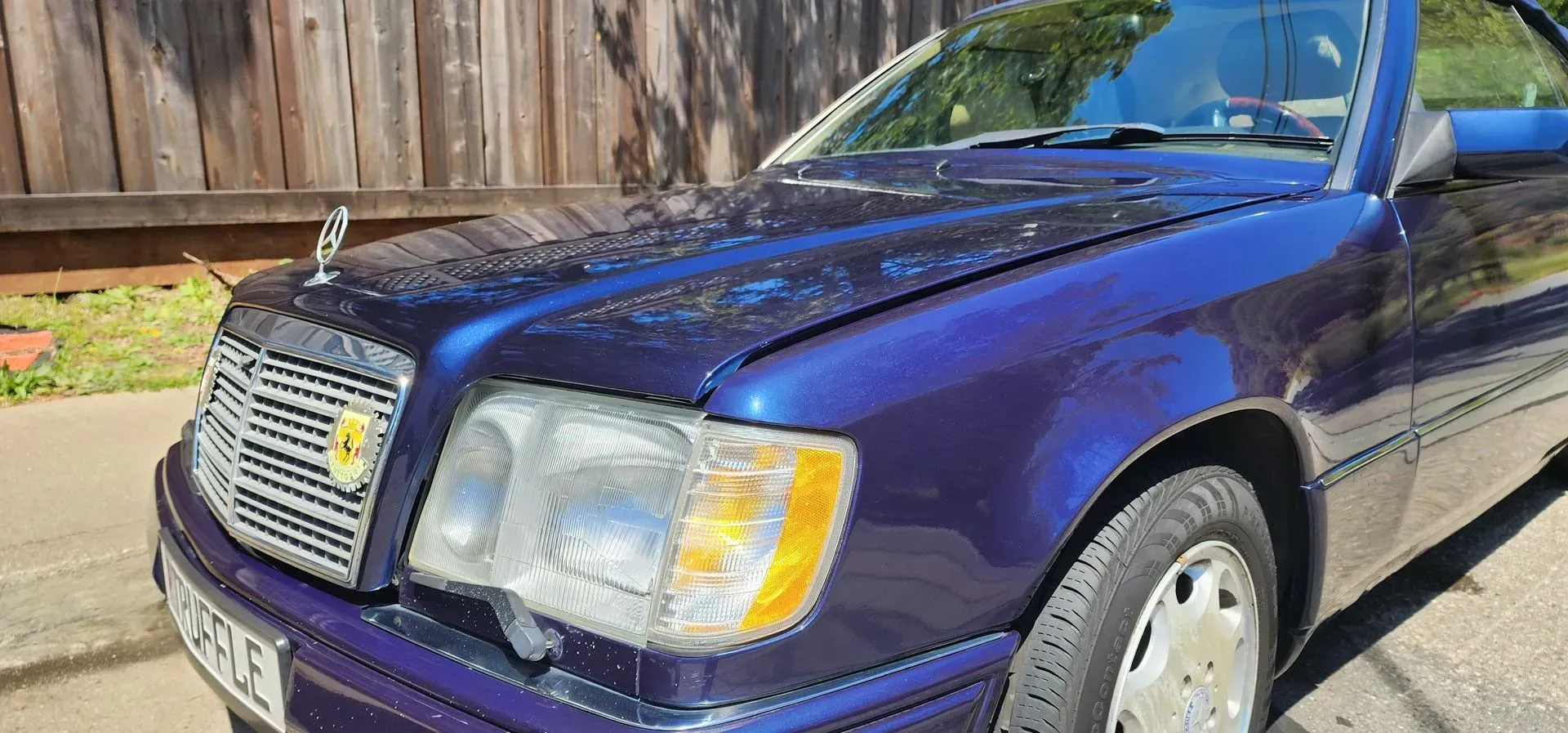 Purple Mercedes sedan parked outdoors in sunlight, front view with wooden fence behind it