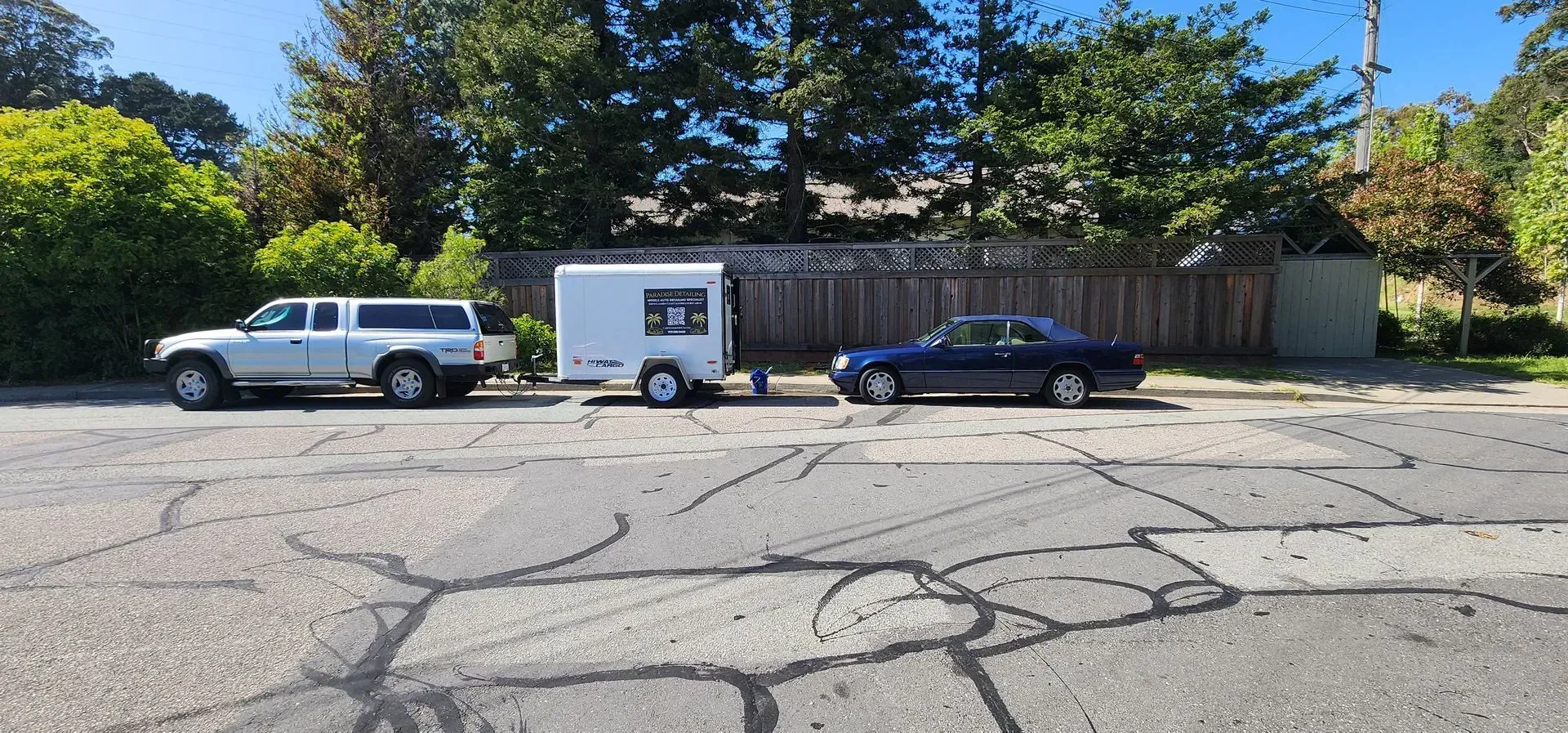 Parked white pickup, small white trailer, and dark sedan along a wooden fence on a cracked street.