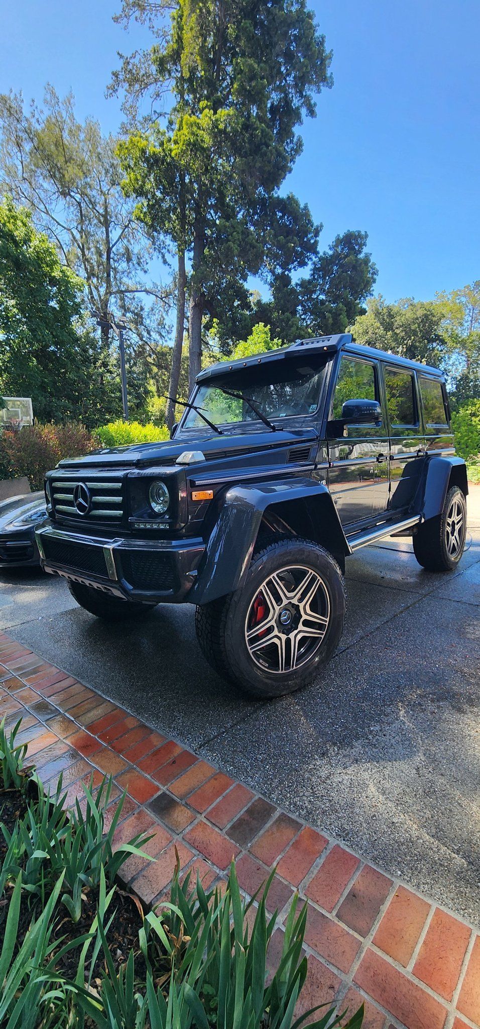Black off-road SUV with roof rack and bull bar parked on gravel beside trees and brick edging