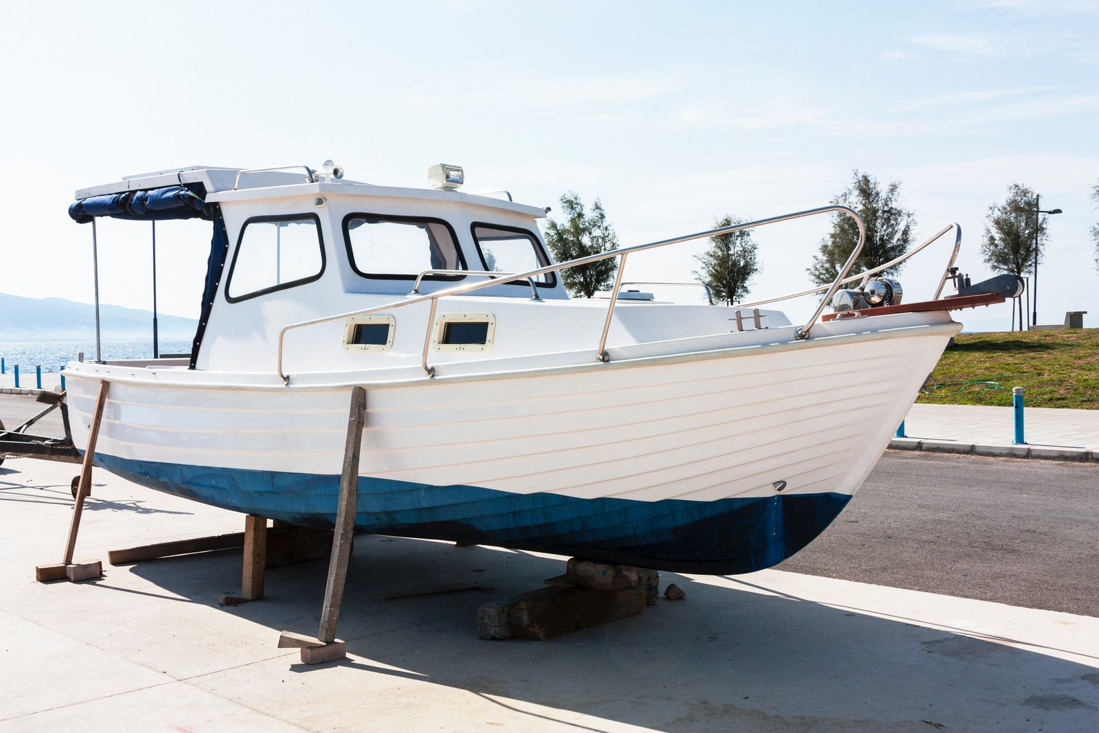 White boat on stands in a boatyard by the water, with a blue hull and open cabin windows.