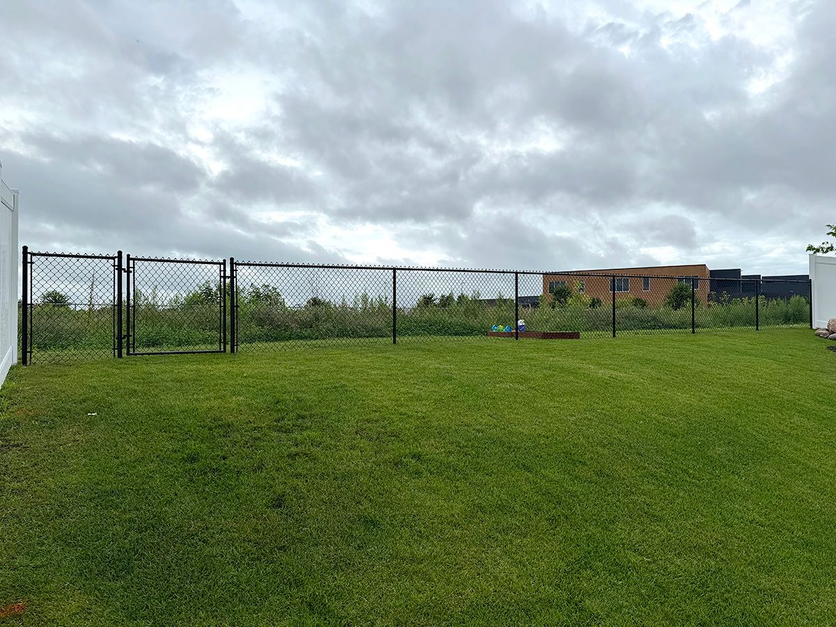 A chain-link fence encloses a green grassy lawn under a cloudy sky, with a building visible in the background.