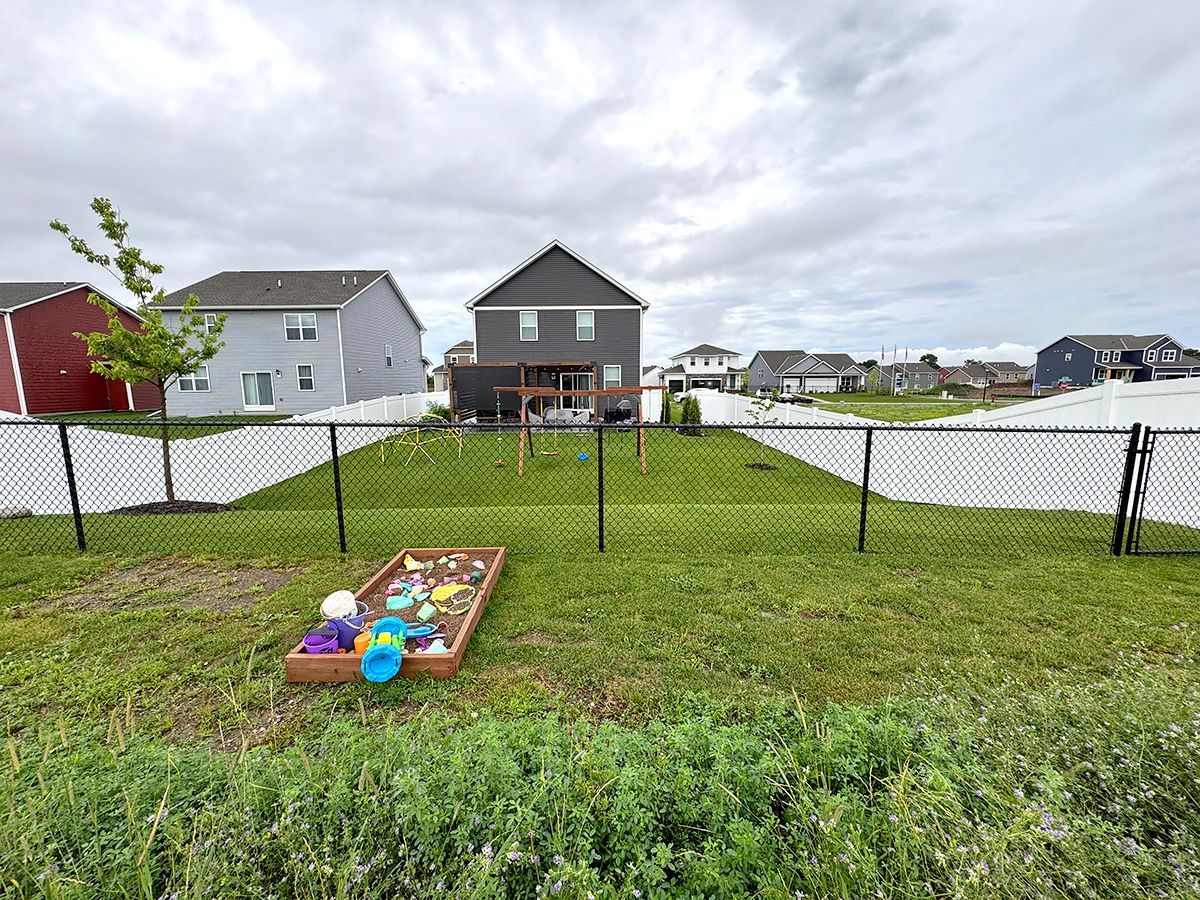 A backyard lawn with a rectangular sandbox in the foreground, a wooden swing set, and suburban houses in the background.