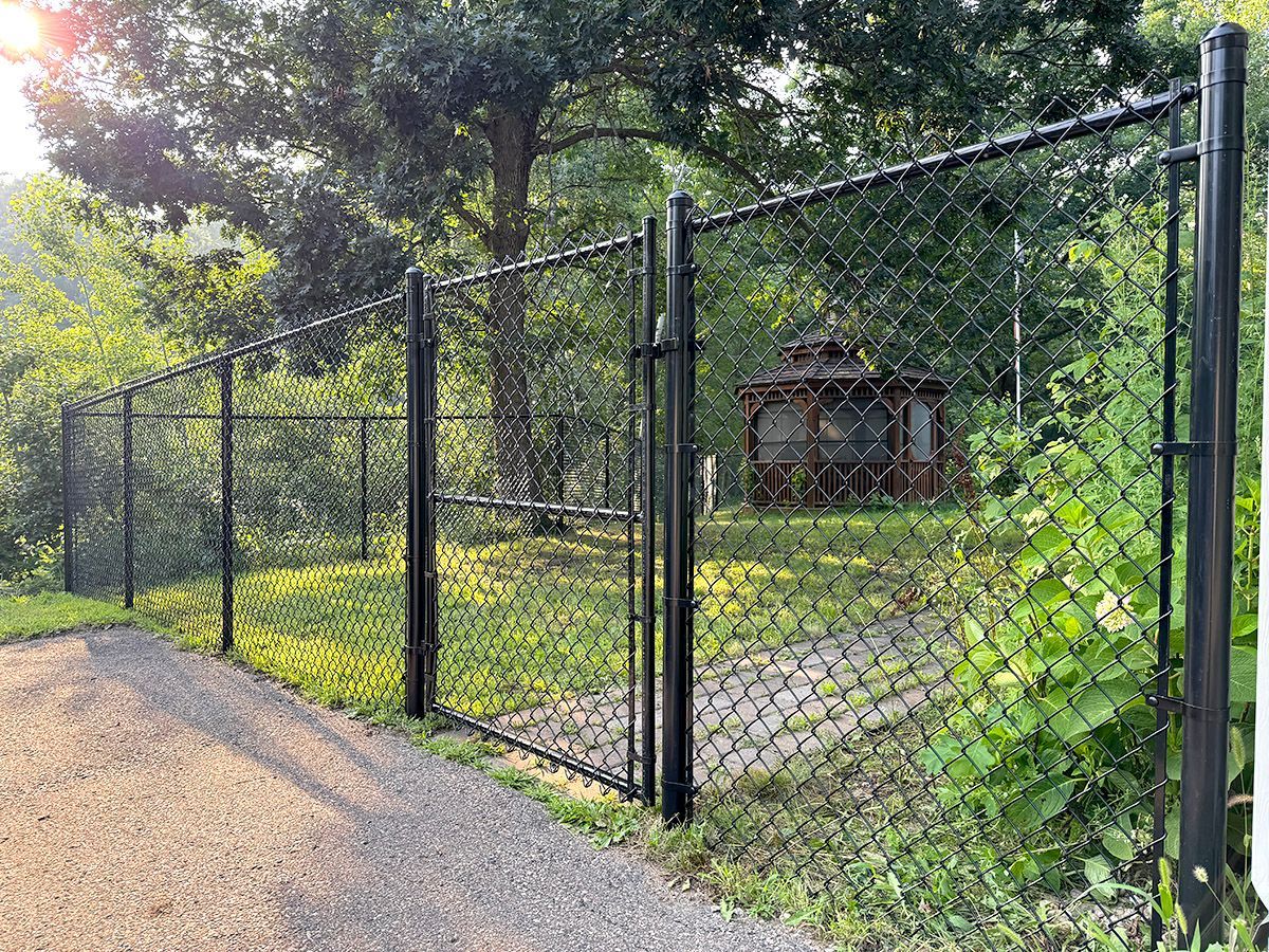 A black chain-link fence with a central gate encloses a grassy yard featuring a wooden gazebo surrounded by trees.