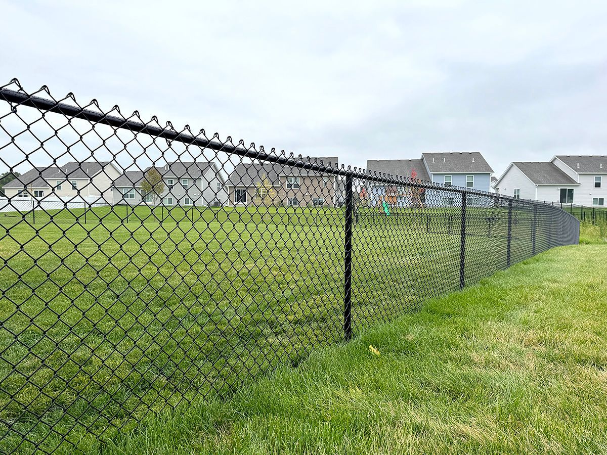 A black chain-link fence curves across a grassy yard in front of several suburban houses under a cloudy sky.