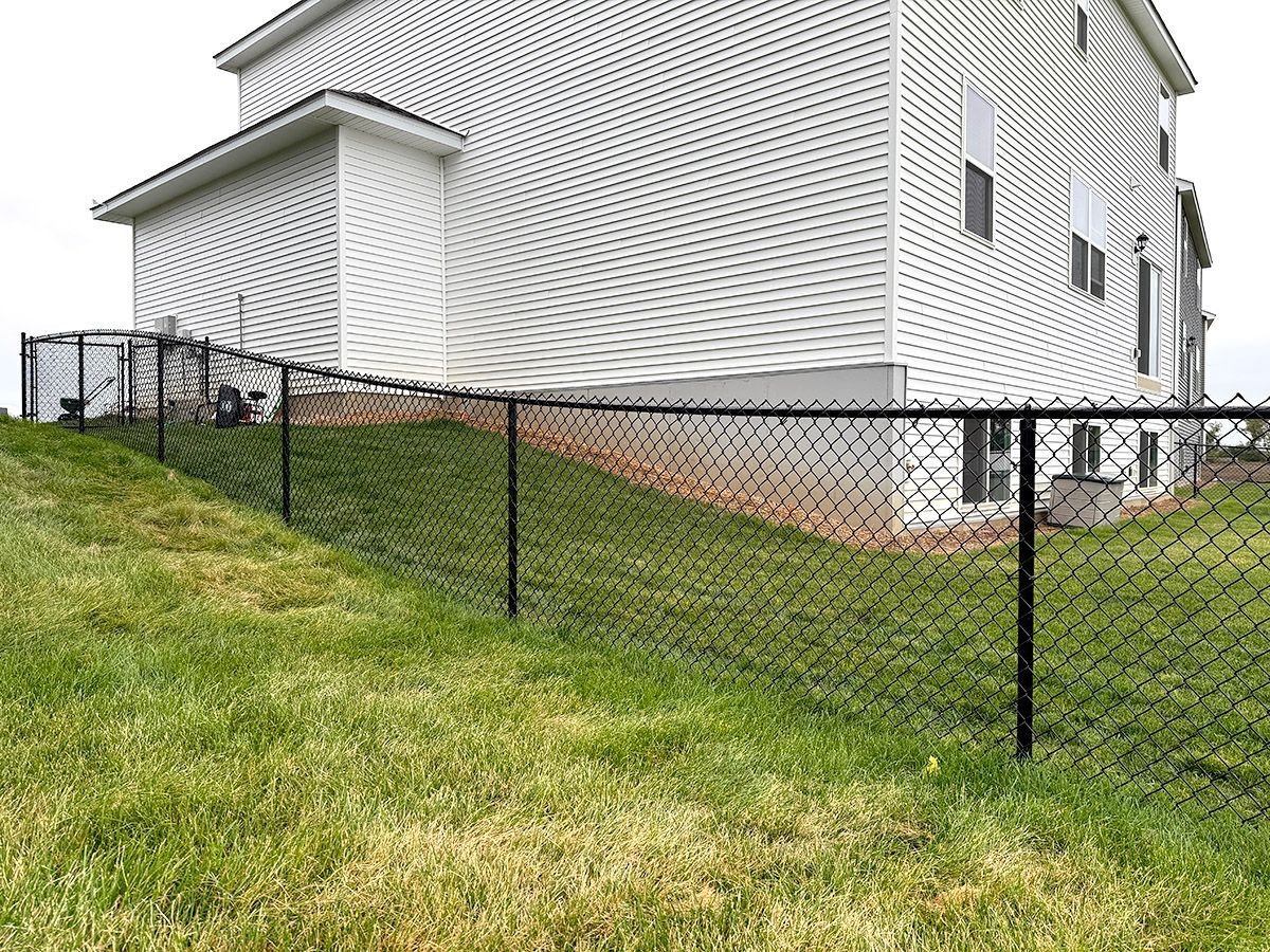 A black chain-link fence runs along a sloping grassy hill next to the side of a light-colored suburban house.