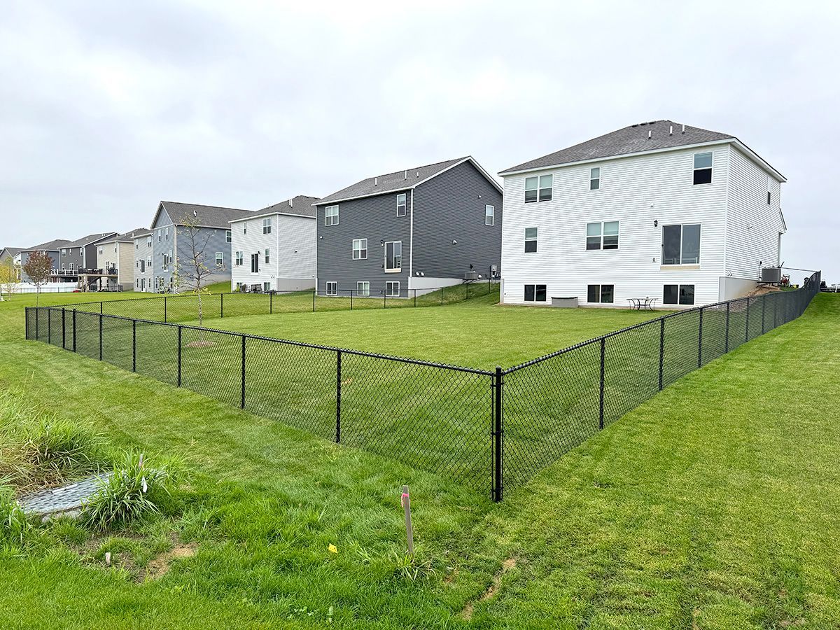 A black chain-link fence encloses a green grassy backyard behind several modern suburban houses on a cloudy day.
