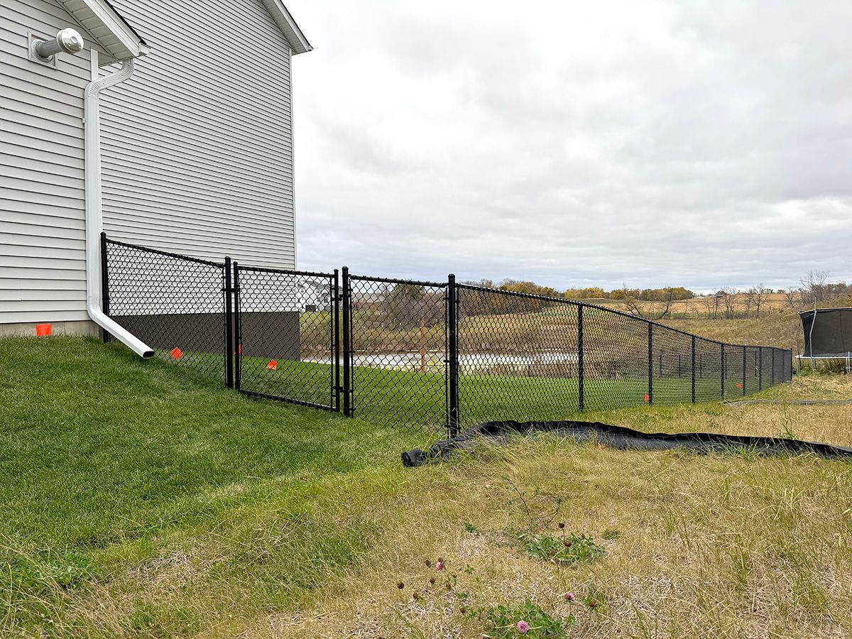 A black chain-link fence with a closed gate extends from the corner of a house across a grassy lawn.