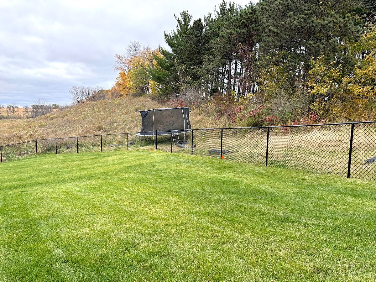 A chain-link fence divides a manicured green lawn from a hilly, grassy field with trees and a trampoline in the distance.