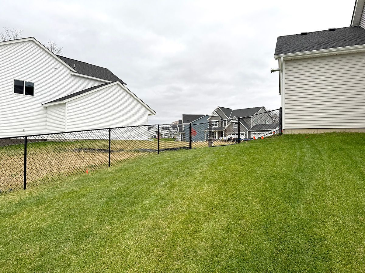 A view from a backyard showing a black chain-link fence separating a grassy slope from neighboring white houses.