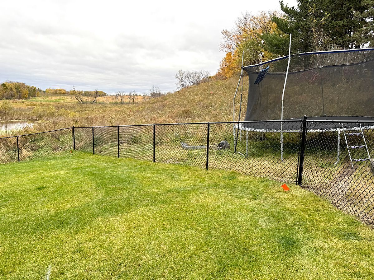 A black chain-link fence lines a green backyard, with a trampoline in the background near a grassy hillside and trees.