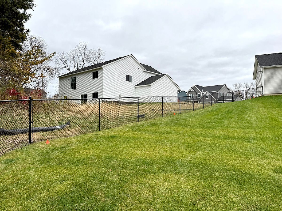 A chain-link fence divides a manicured green lawn from a dry, grassy field behind two suburban houses under a cloudy sky.