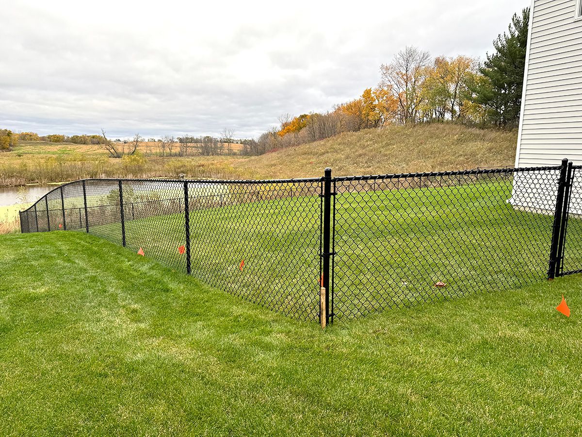 A black chain-link fence encloses a green grassy lawn in a backyard, with a pond and hilly landscape in the distance.