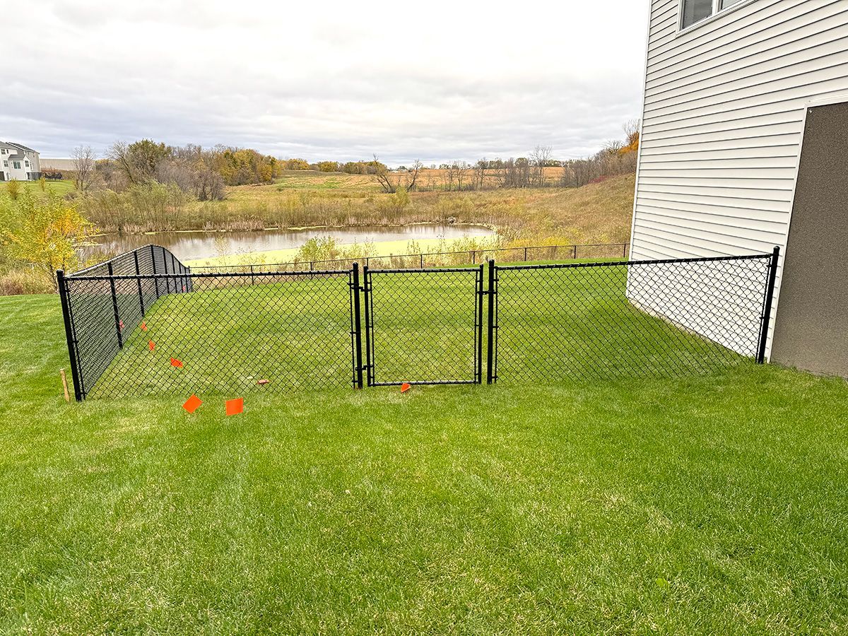 A black chain-link fence encloses a backyard lawn next to a house, with a pond and field in the background.