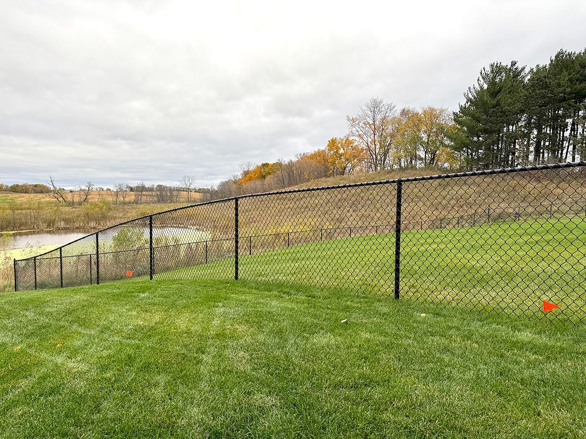 A black chain-link fence divides a lush green lawn from a pond and a golden, autumn-colored hillside under a cloudy sky.