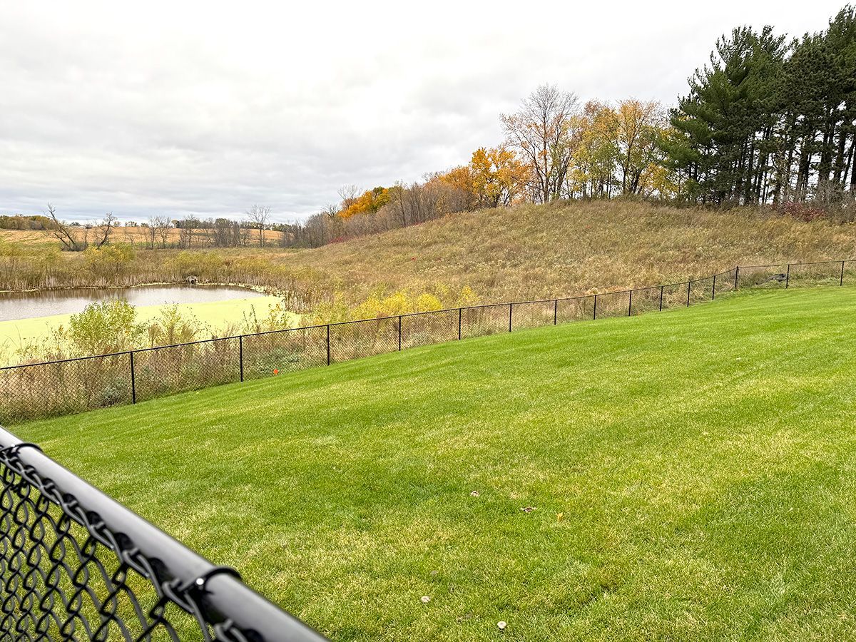 A green lawn slopes down toward a chain-link fence, a pond, and a wooded hill under a cloudy autumn sky.