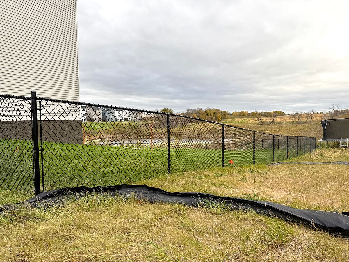 A black chain-link fence runs along a sloping grassy yard next to a building, with a field and pond in the distance.