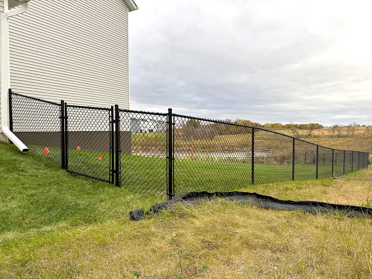 A black chain-link fence extends from the corner of a beige house across a grassy yard toward an open field.