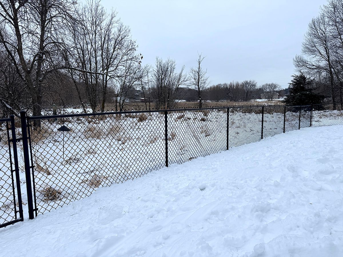 A black chain-link fence encloses a snow-covered yard in front of a field and bare trees on a winter day.
