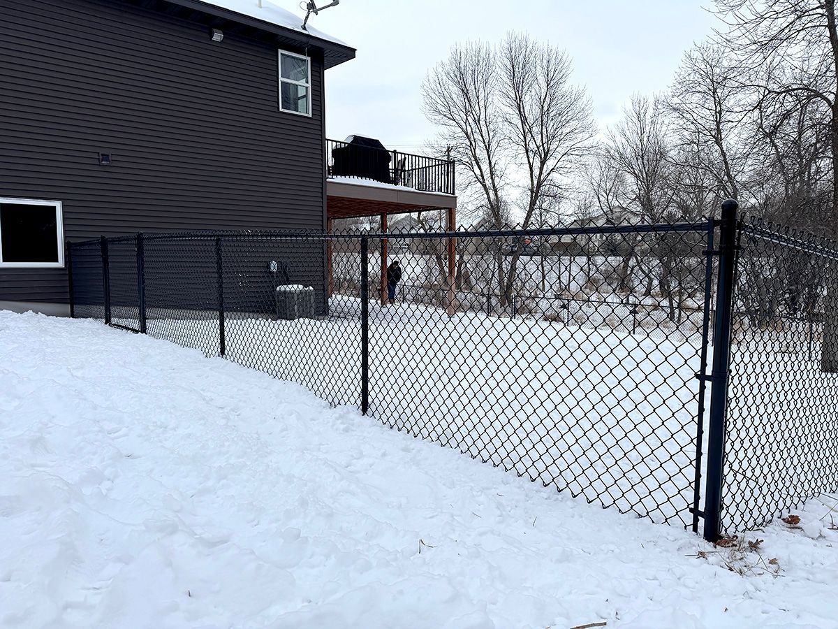 A black chain-link fence surrounds a snowy backyard next to a dark-sided house with a raised wooden deck.