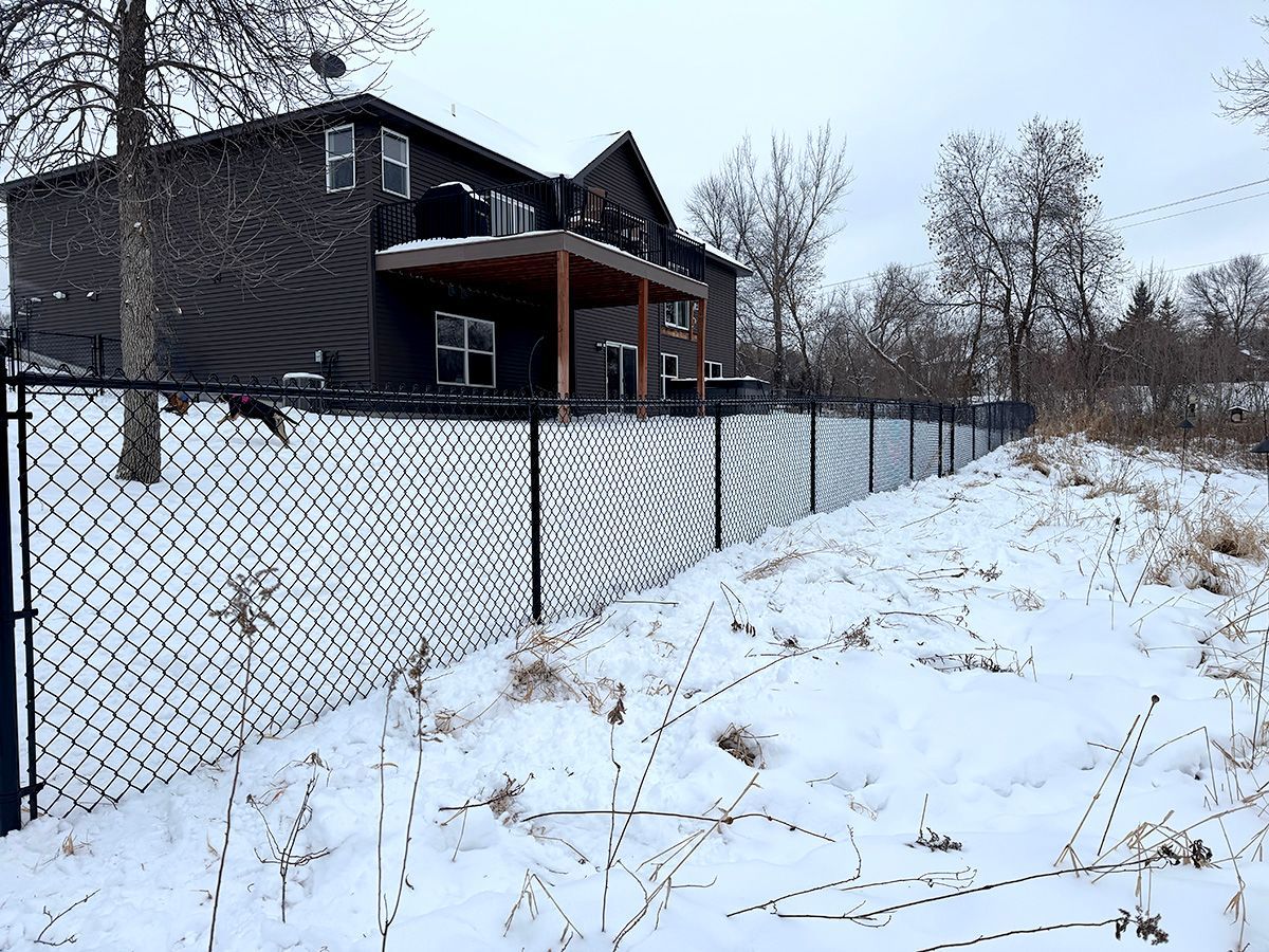 A dark multi-story house with a wooden deck stands behind a black chain-link fence with white privacy slats in the snow.