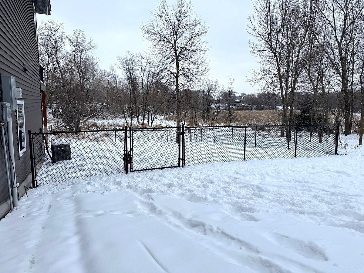 A fenced yard area behind a house covered in snow, with bare trees in the background under a cloudy sky.