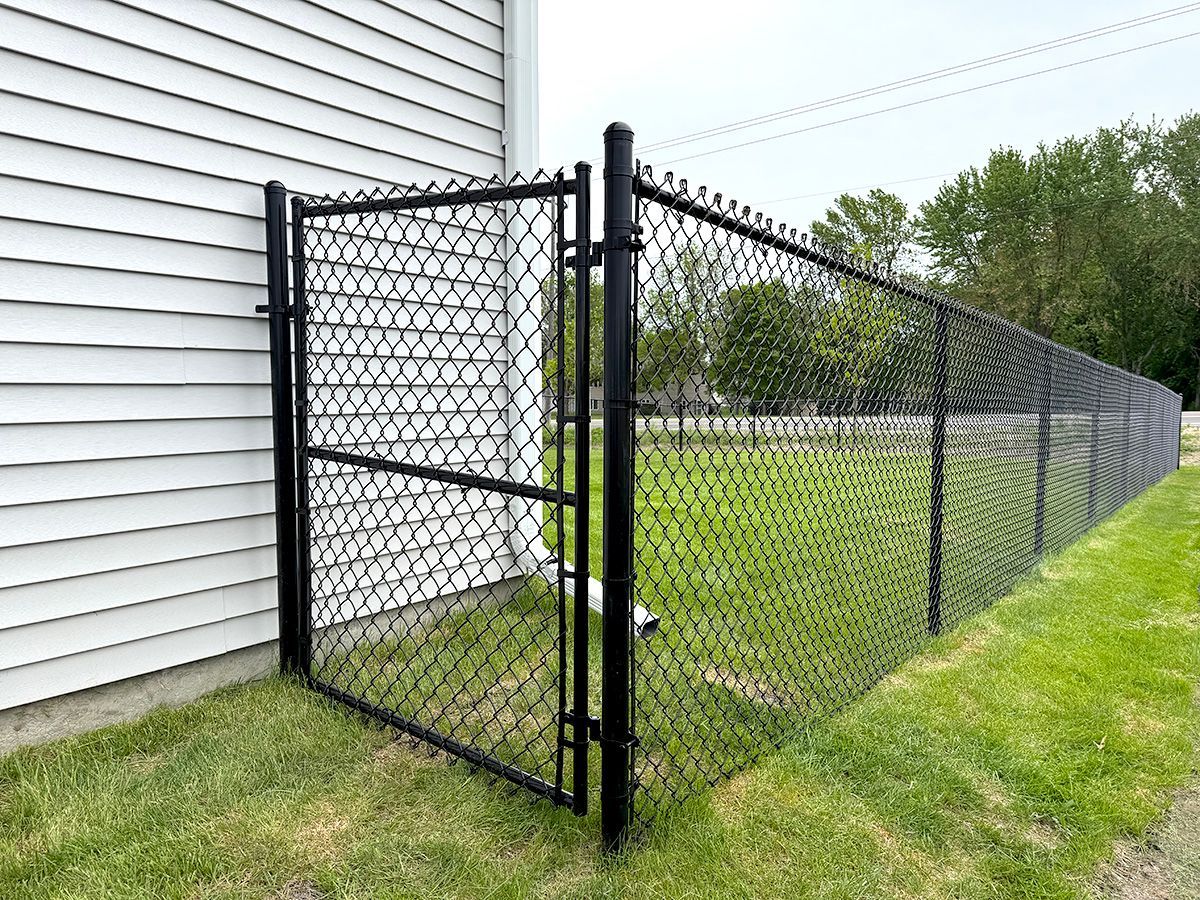 A black chain-link fence and gate connected to the side of a white-sided house with a grassy lawn in the background.