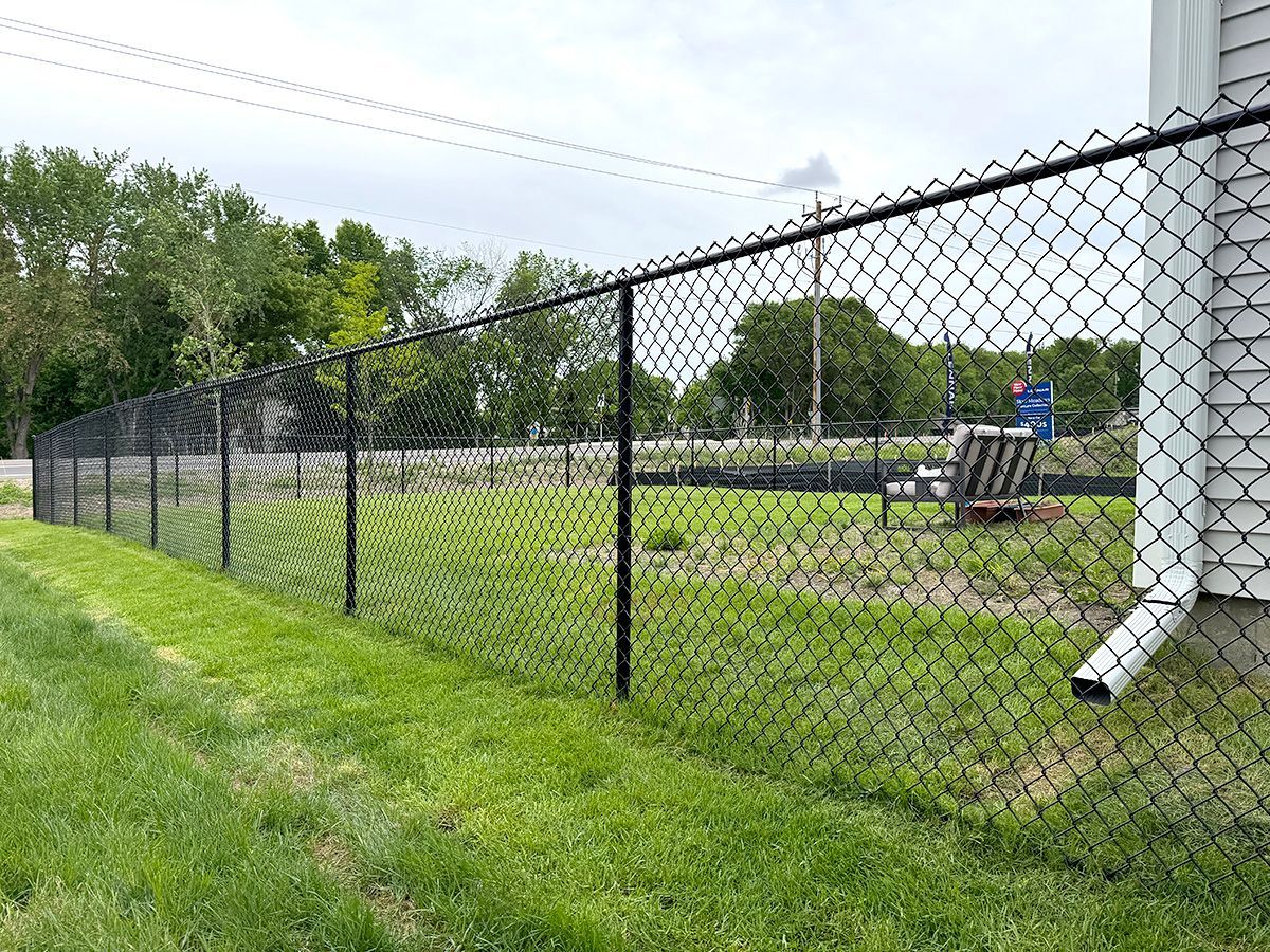 A black chain-link fence encloses a green grassy backyard next to the corner of a house with a white downspout.