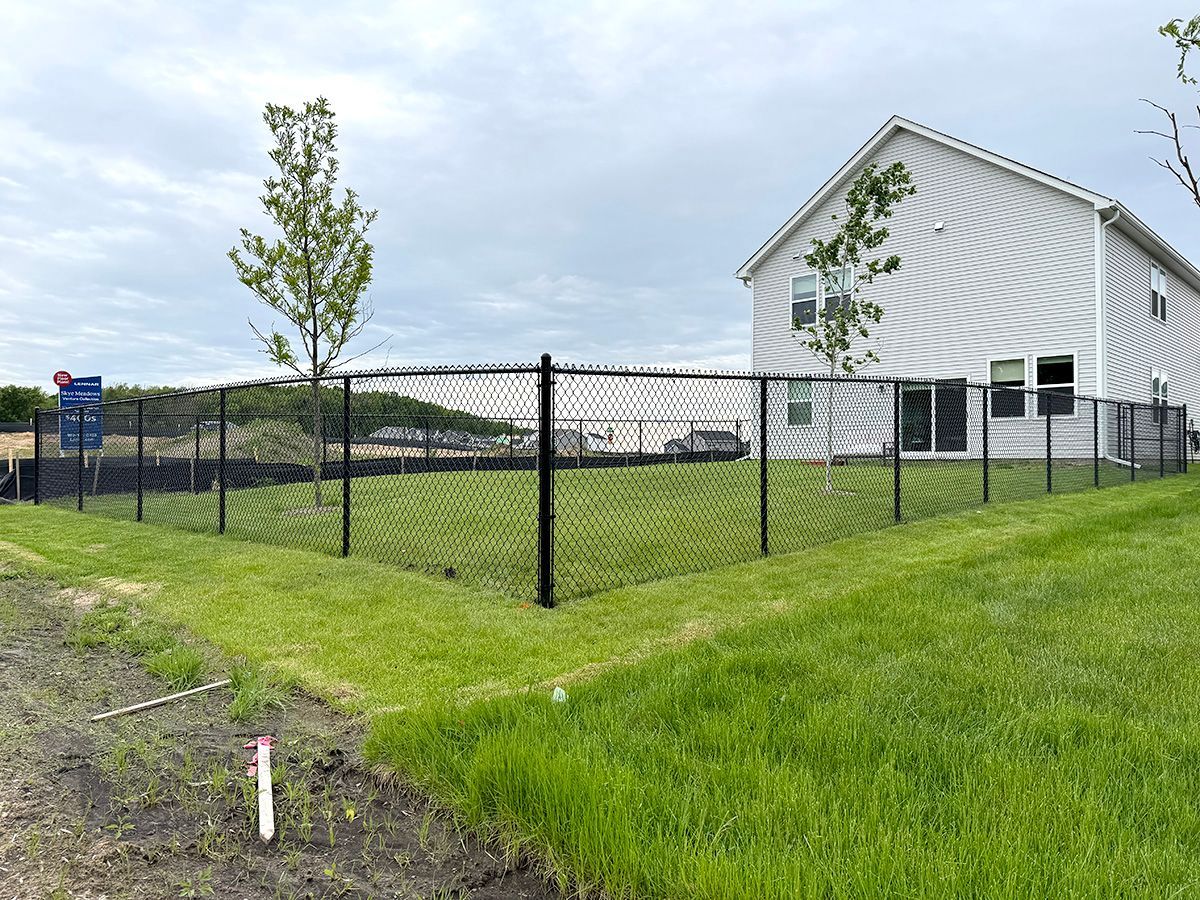 A black chain-link fence surrounds the side and back yard of a two-story light gray house on a grassy residential lot.