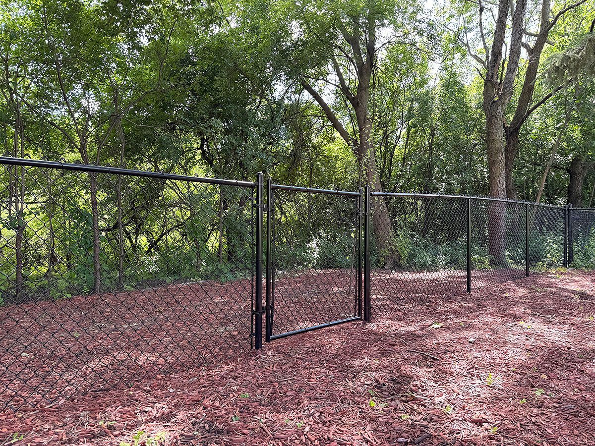 Black chain-link fence with a central gate installed in a mulched area with a wooded background.