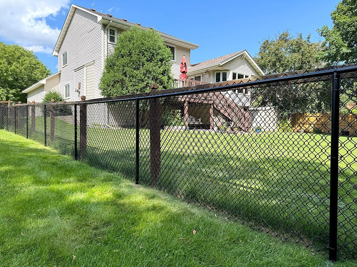 A black chain-link fence runs along a green residential lawn toward a two-story beige house with a wooden deck.