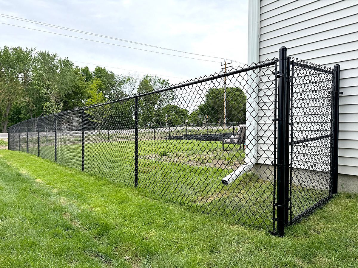 A black chain-link fence runs along the side of a white house, bordering a grassy yard with trees in the background.