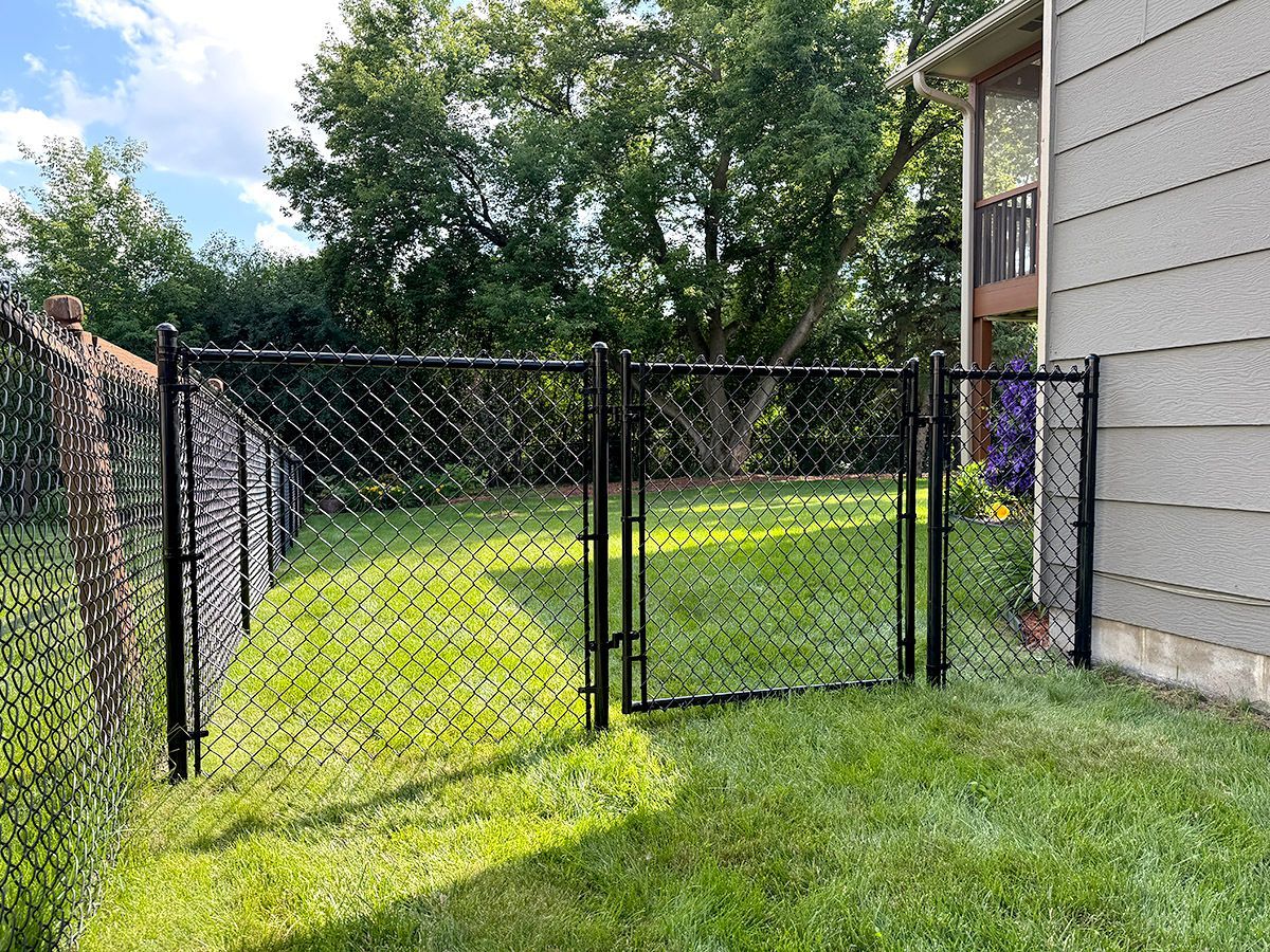 A black chain-link fence with a gate stands on a grassy lawn next to the side of a grey house.