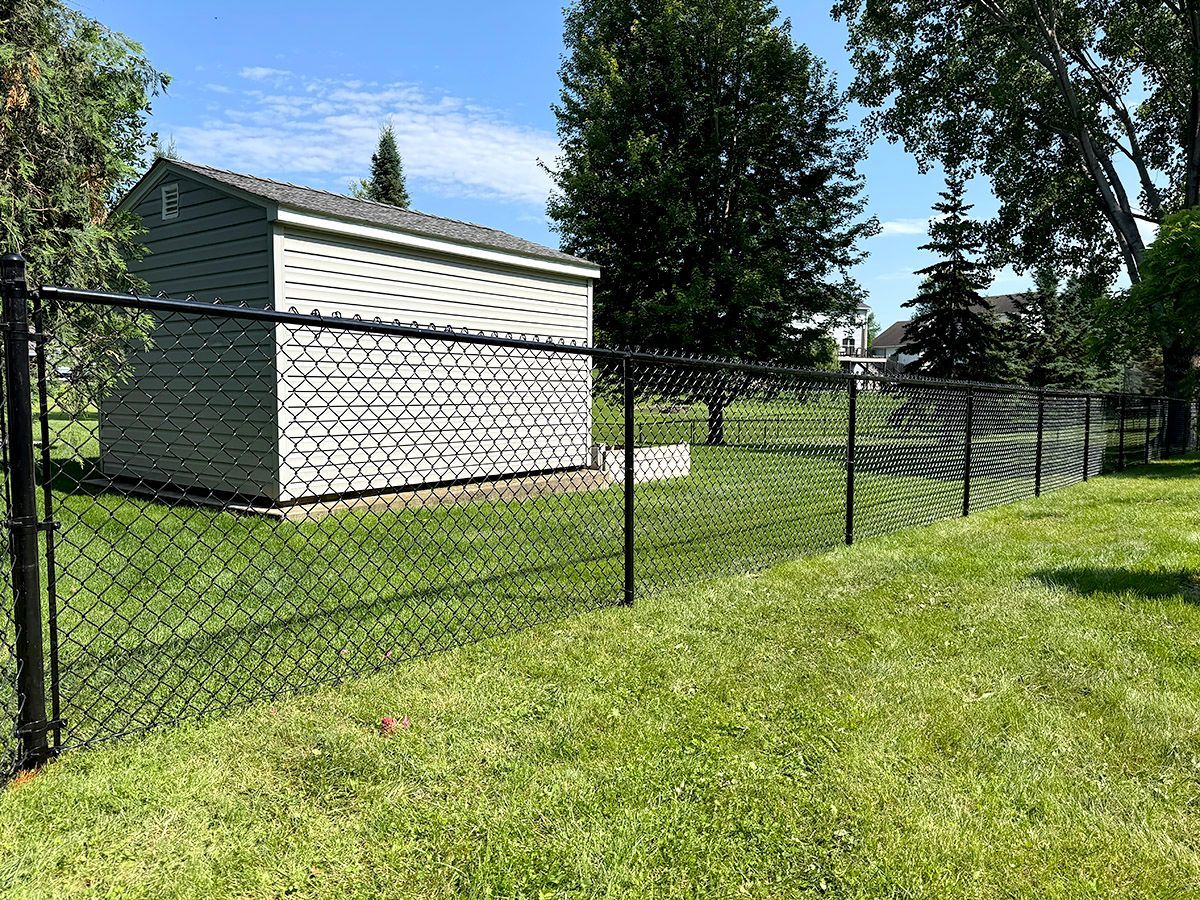A black chain-link fence runs along a green lawn in front of a gray storage shed on a sunny day.