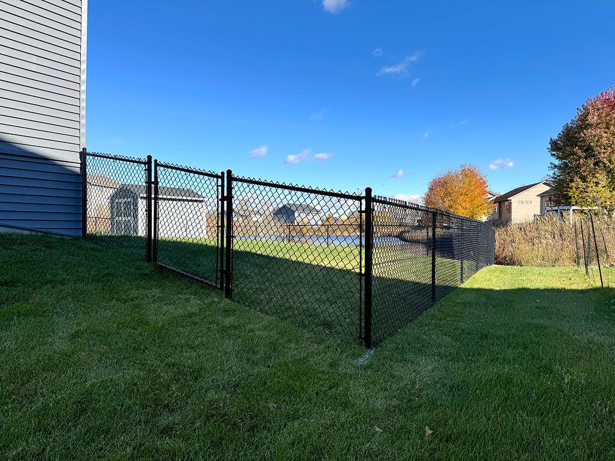 A black chain-link fence runs along a sloping grassy backyard, with a shed and pond visible in the distance under blue sky.