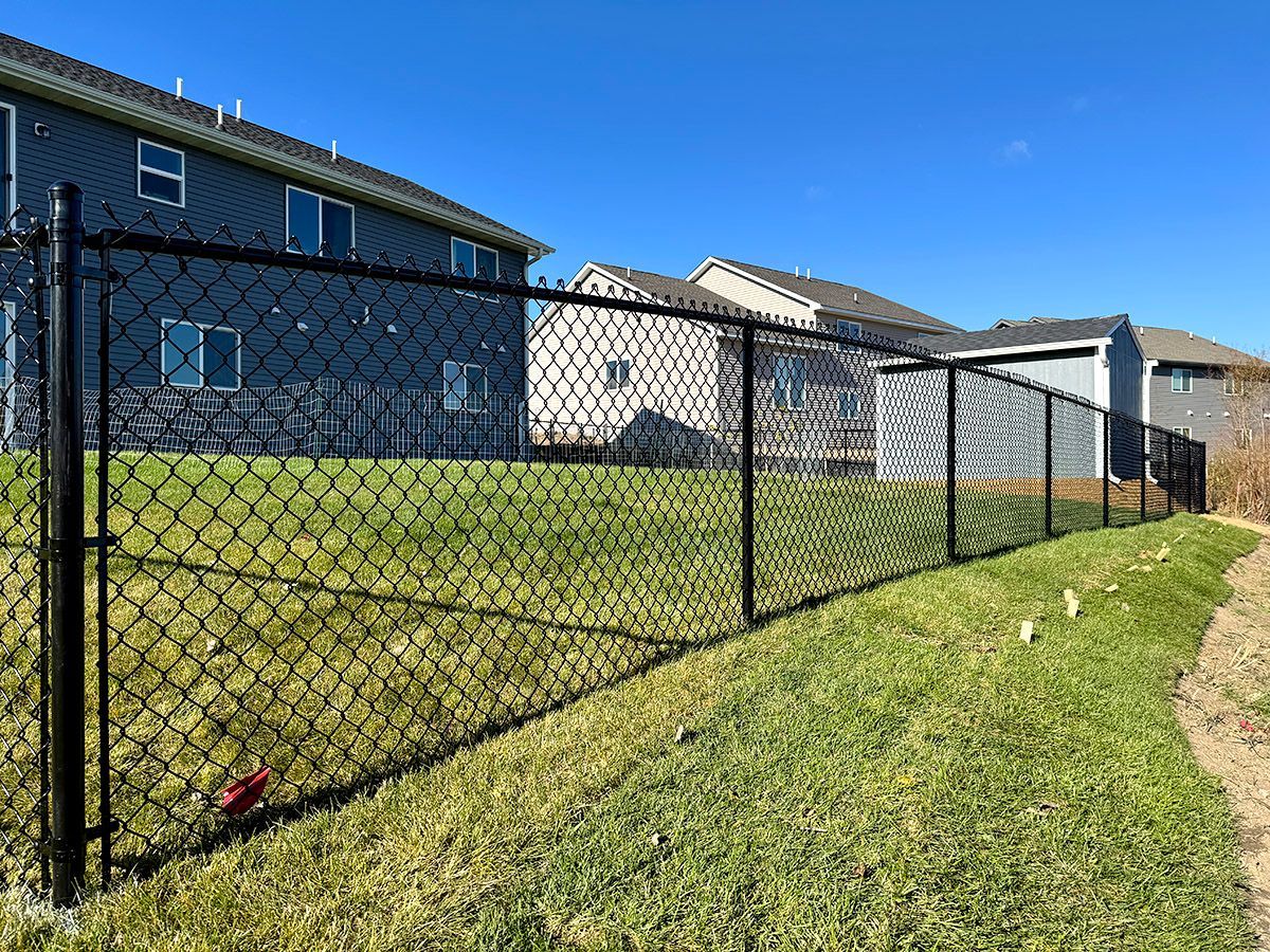A black chain-link fence runs along a sloping grassy yard in front of several suburban homes under a clear blue sky.