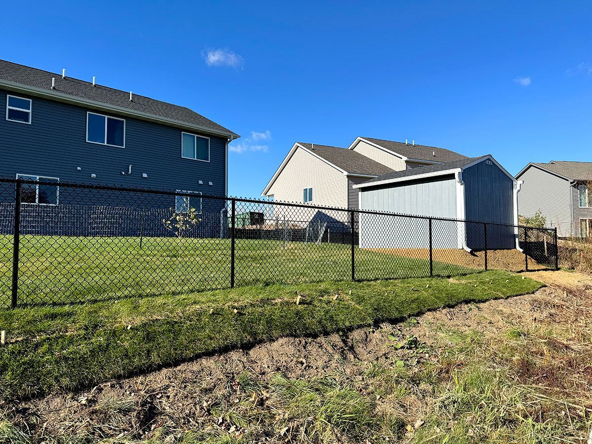 A blue suburban house next to a light-colored home with a gray storage shed, all enclosed by a black chain-link fence.