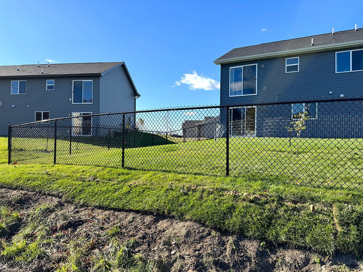 Two houses with blue-gray siding and black chain-link fences sit on a grassy hill under a bright blue sky.