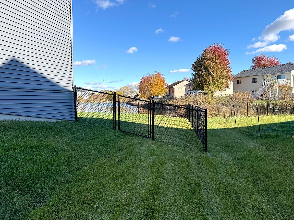 A black chain-link fence extends from the side of a light gray house toward a pond on a sunny day.