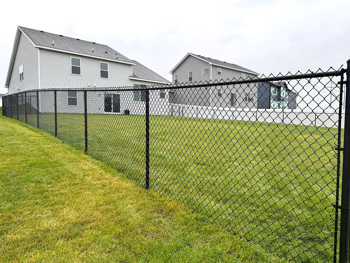 A black chain-link fence encloses a green grassy backyard behind two suburban houses under a cloudy sky.