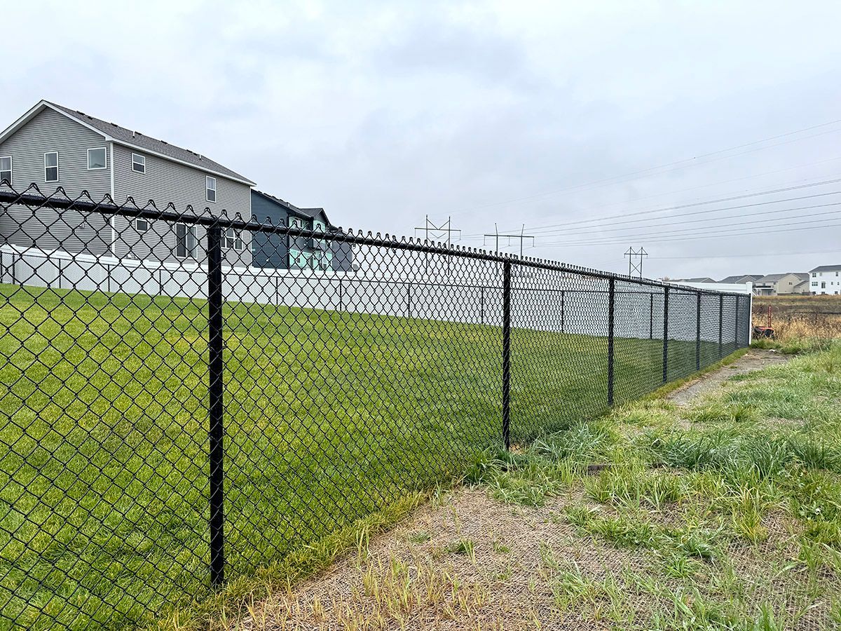 A black chain-link fence lines a grassy slope in front of residential houses under an overcast sky.